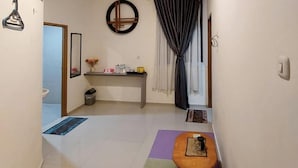 A small, clean room with a purple mat, a wooden bench, a small table, a mirror, a shelf with toiletries, and a window with curtains. - TwoSpaces Living at Maximus Inn (Palembang)