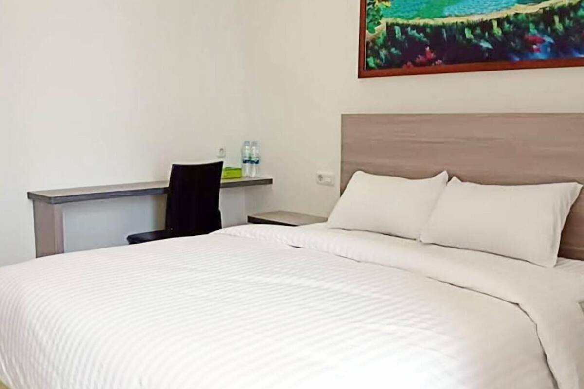 A hotel room with a large bed, a desk, and a chair. There is a painting on the wall and a shelf with water bottles.