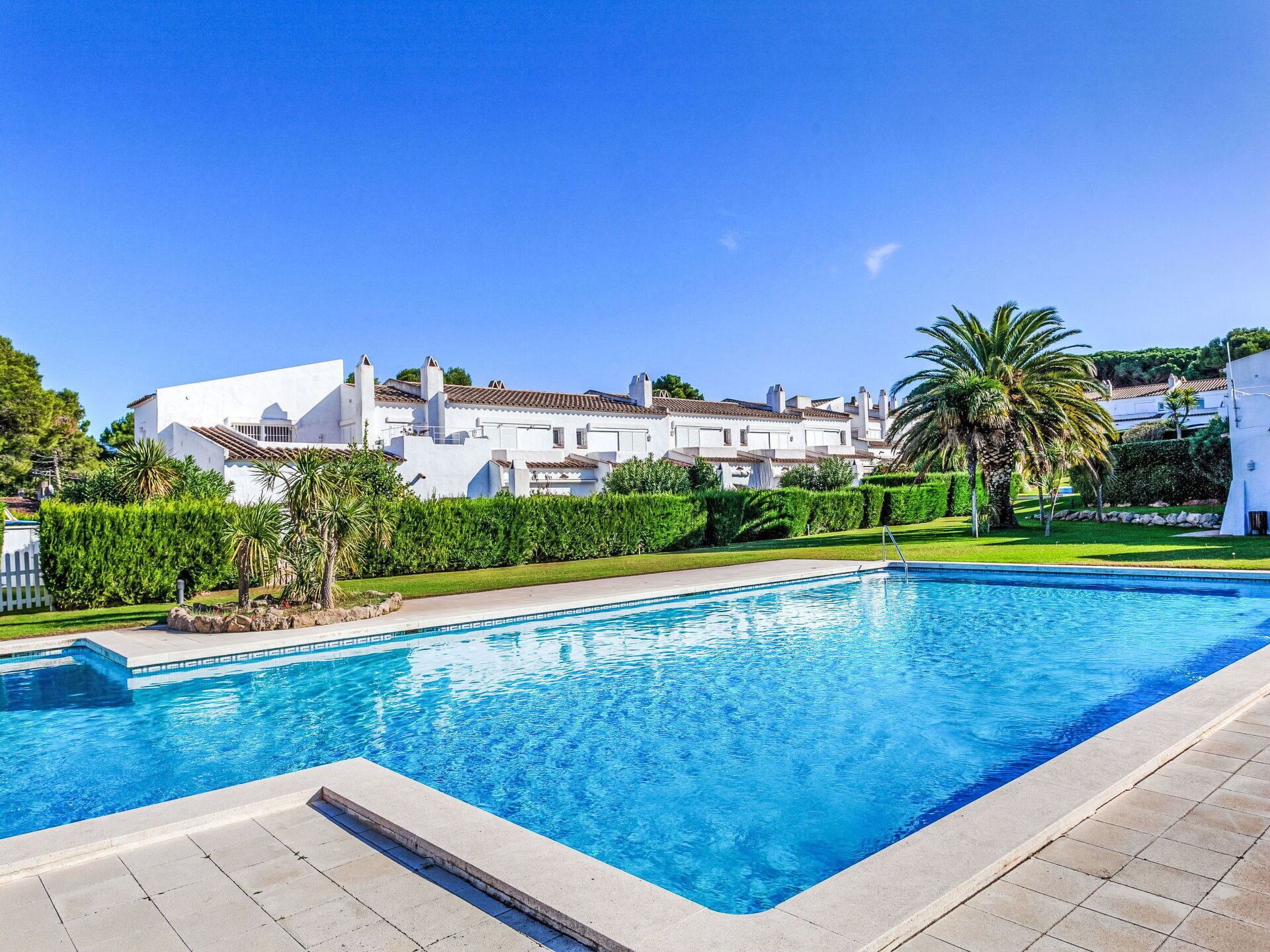 A swimming pool with a clear blue water surface, surrounded by a paved deck. In the background, there are white buildings with red-tiled roofs and a well-maintained lawn with palm trees.