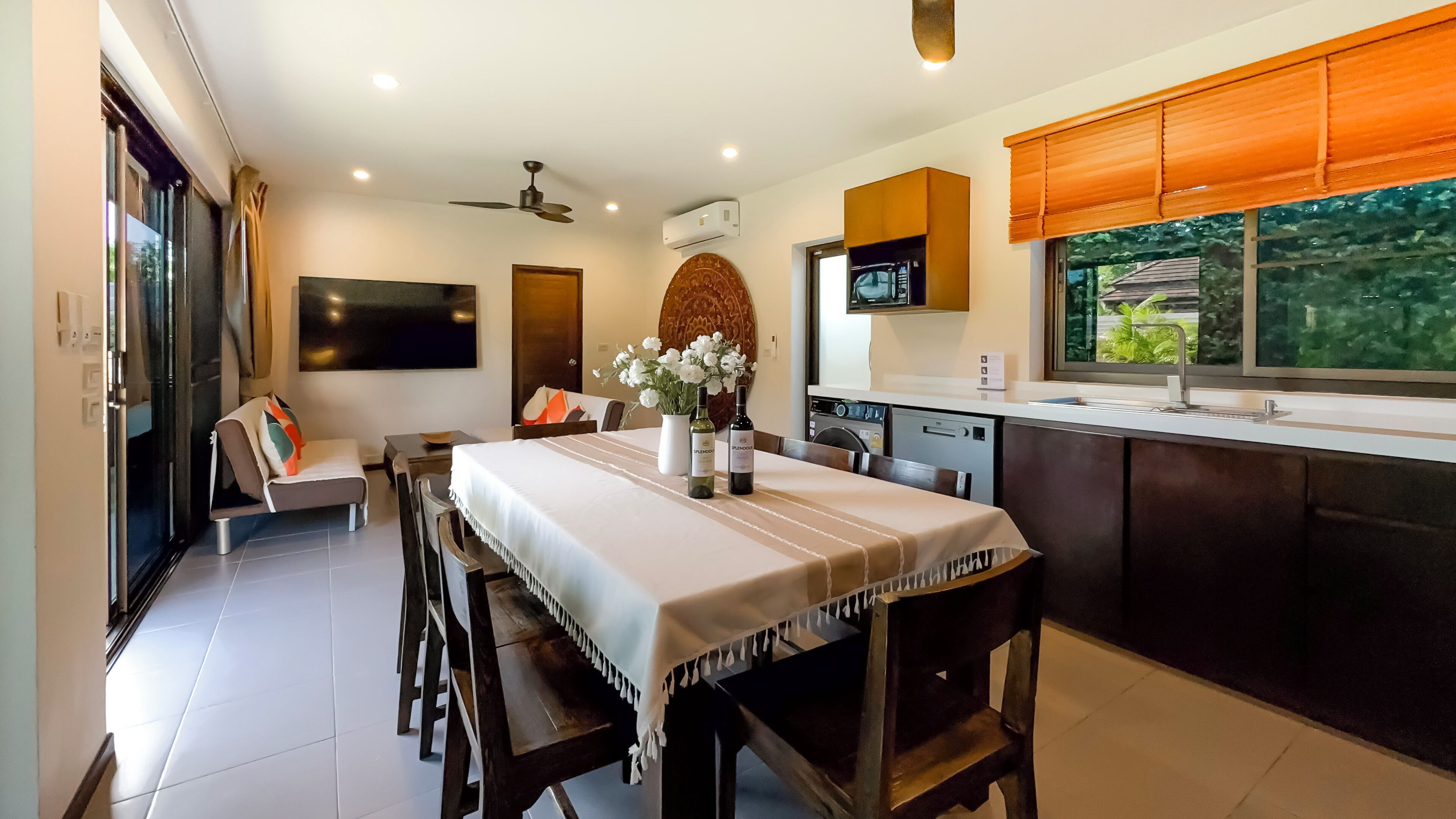 A modern kitchen with a dining area, featuring a large table, chairs, a TV, and a window with a view of greenery.