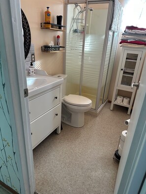 A compact bathroom with a white toilet, a sink, and a shower enclosure. There are shelves with toiletries and a cabinet with towels. - Cozy cabin - Highlands of Belair (Bélair)