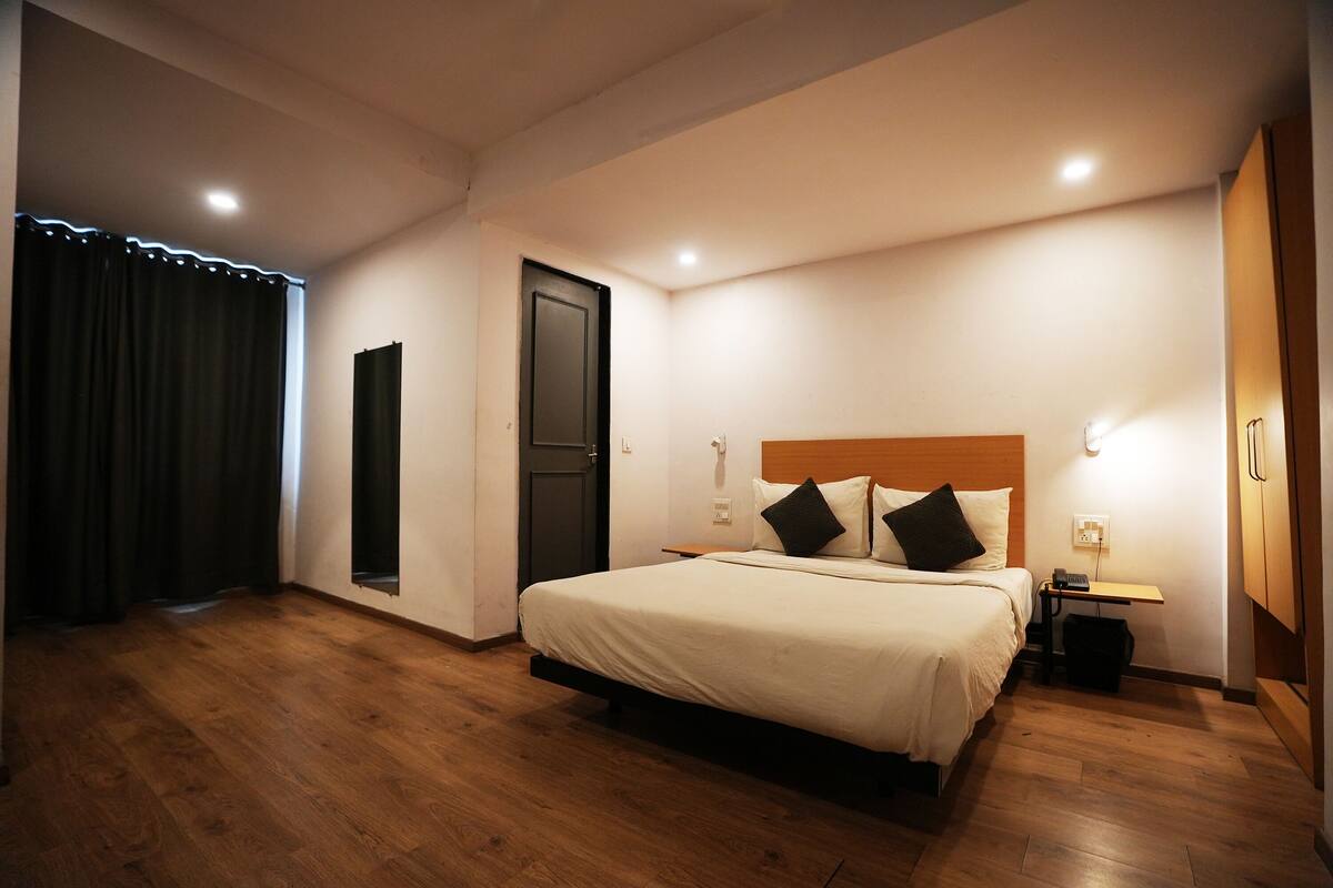 A hotel room with a large bed, a wooden floor, a door, a nightstand with a lamp, and a window with curtains.