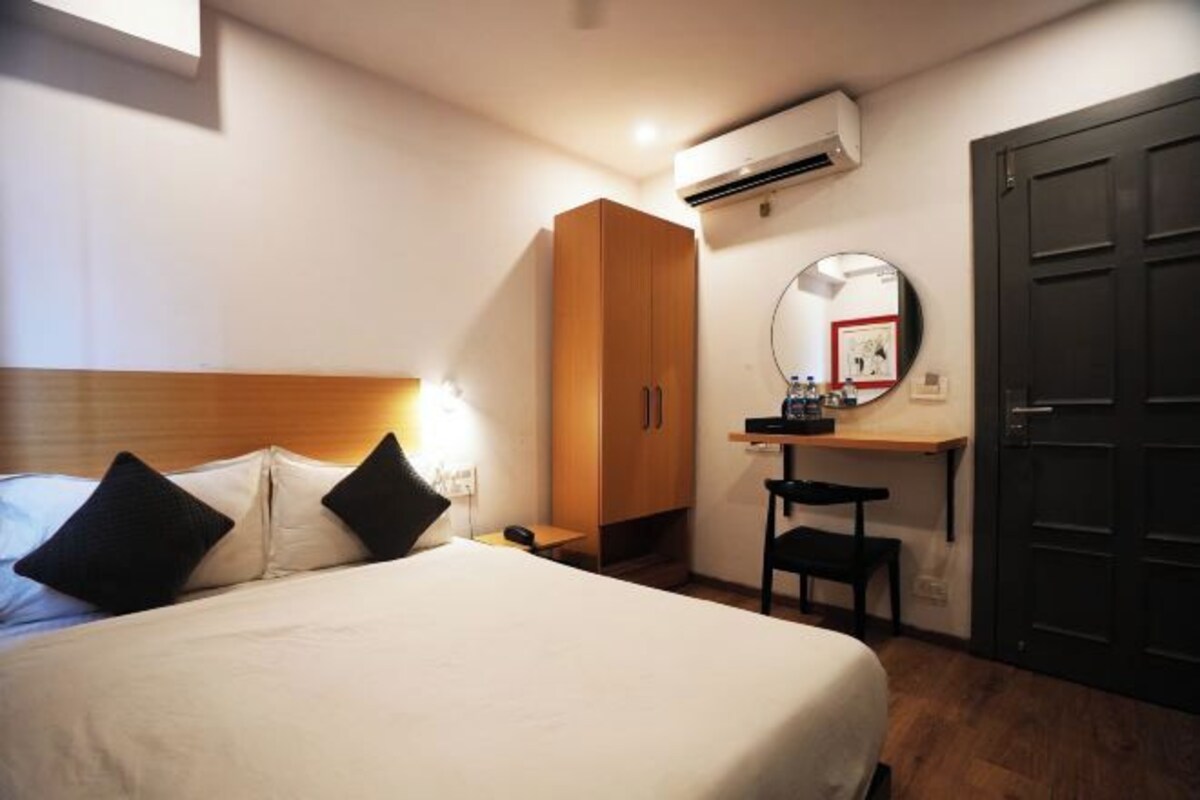 Ein Hotelzimmer mit einem Bett, einem Holzschrank, einem Schreibtisch mit Stuhl, einem runden Spiegel und einer dunklen Tür.