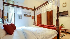 Una cama con dosel con ropa de cama blanca y almohadas rojas, un cabecero de madera y un espejo encima. Hay un televisor en la pared a la derecha y una planta sobre una mesita de noche.