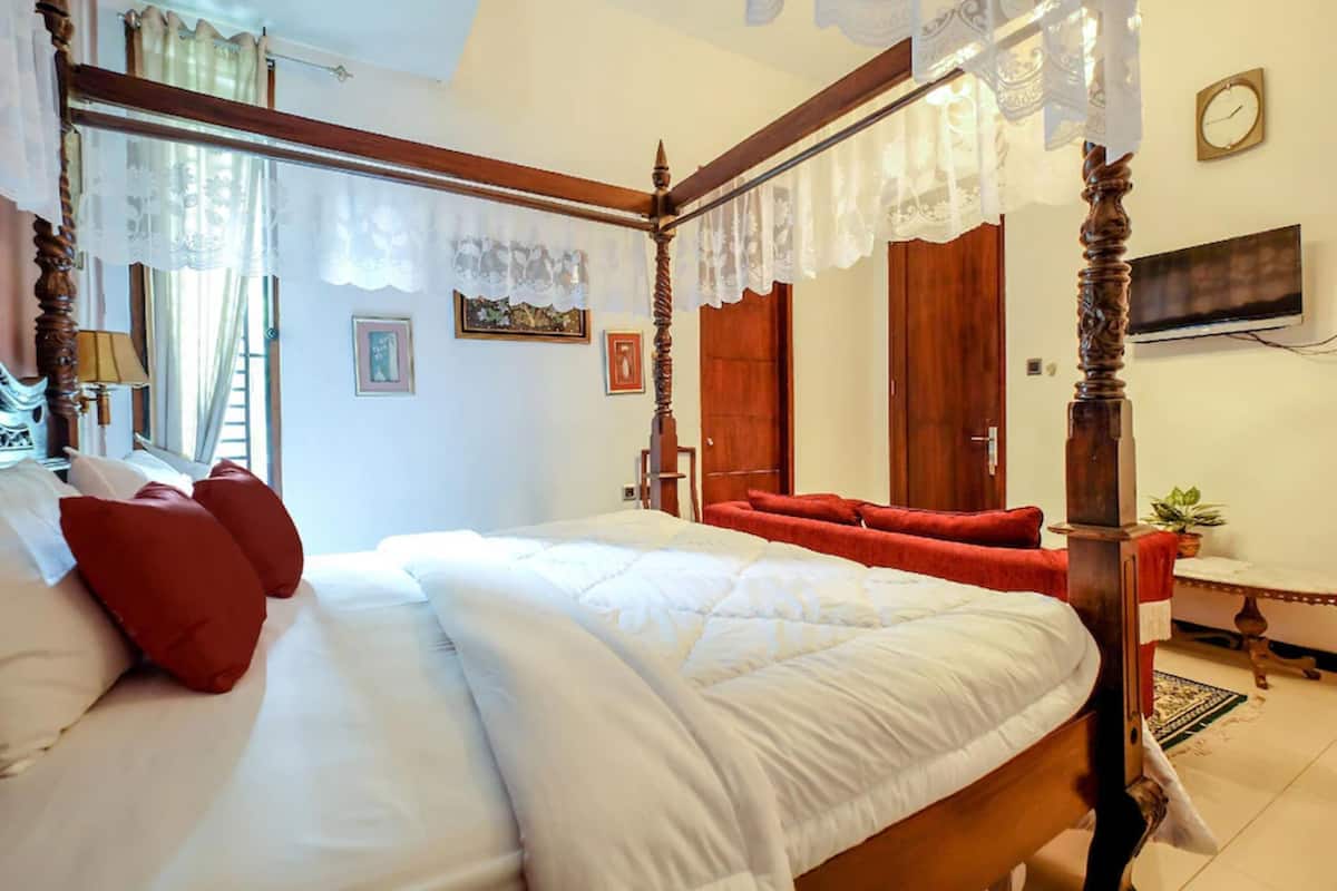 A four-poster bed with white bedding and red pillows, a wooden headboard, and a mirror above it. There is a television on the wall to the right and a plant on a small table beside the bed.
