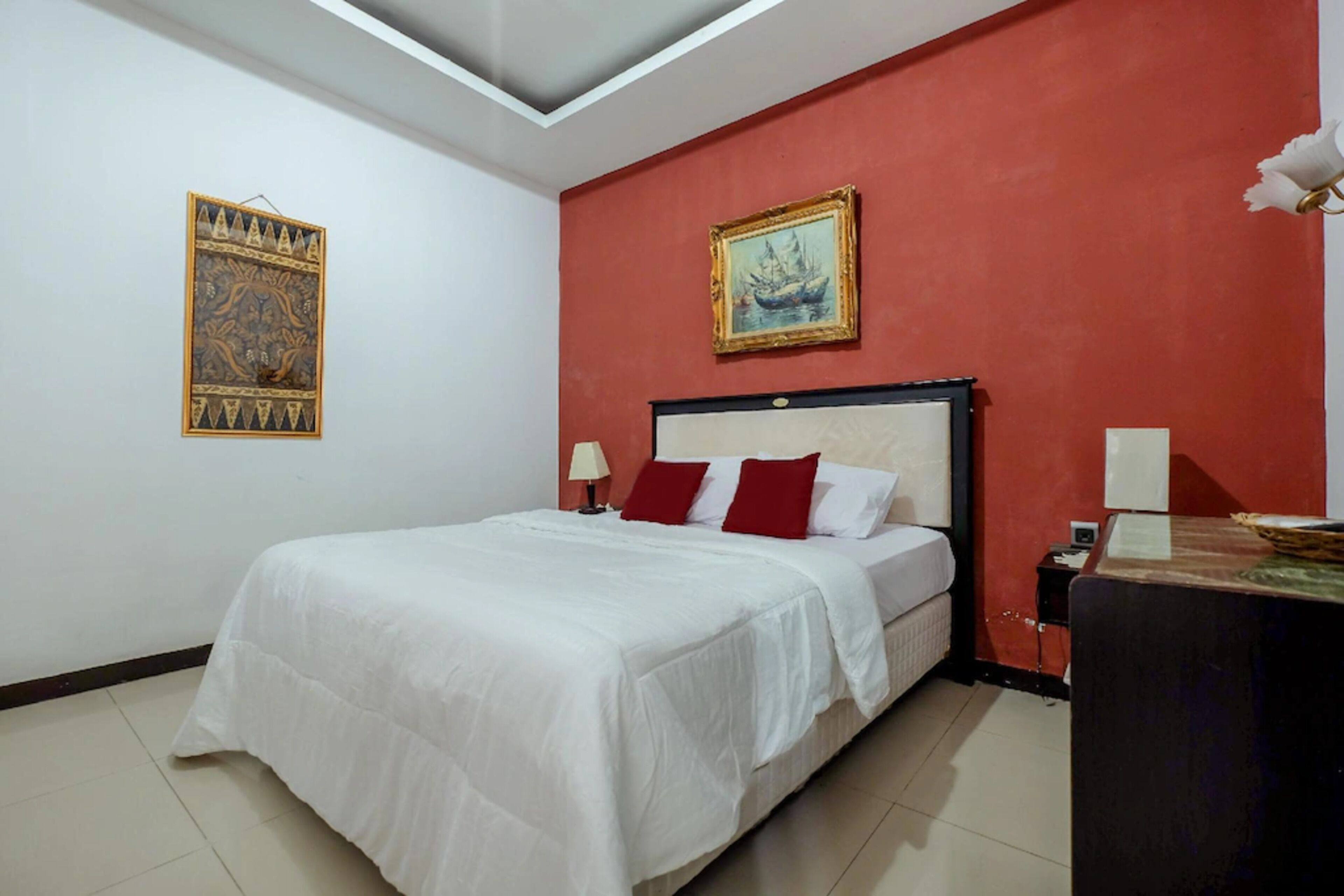 A bedroom with a bed, a nightstand, a desk, a red wall, and two framed pictures.