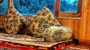 Una habitación acogedora con techo de madera, un ventanal con vistas a un bosque y una cama con almohadas estampadas con motivos florales.