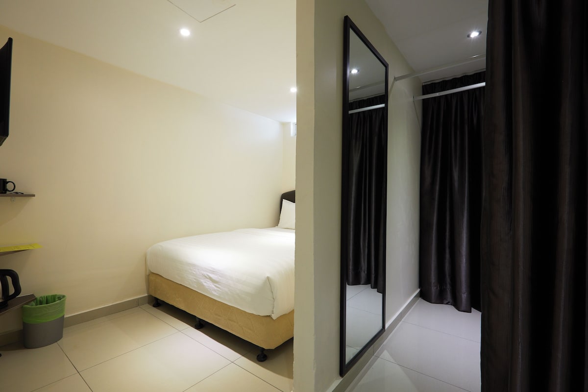 A hotel room with a bed, a mirror, a curtain, and a trash bin.