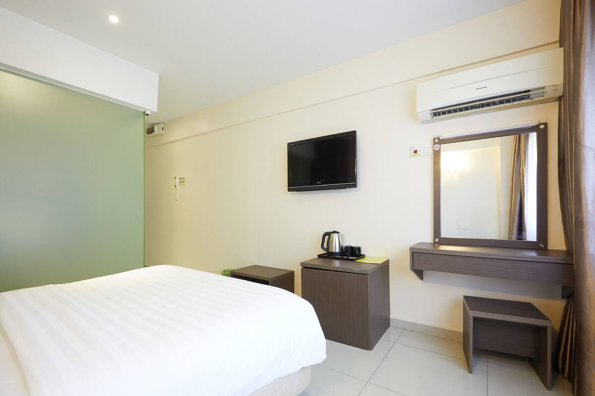 A hotel room with a bed, a small desk with a kettle, a flat-screen TV, and a mirror.