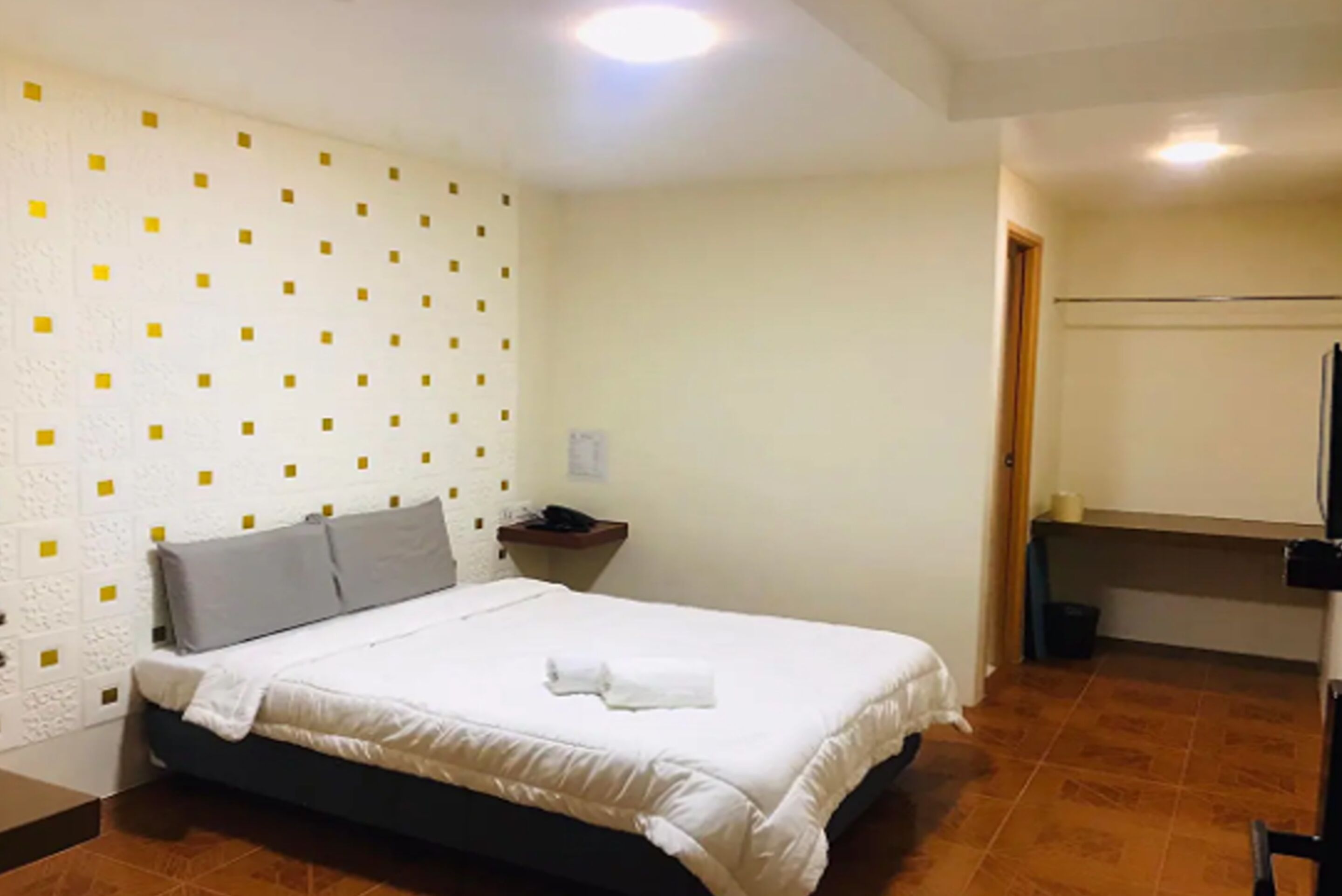 Ein ordentlich bezogenes Bett mit weißer Decke und grauen Kissen, ein kleiner Nachttisch mit Telefon und Lampe sowie eine Wand mit geometrischem Muster.