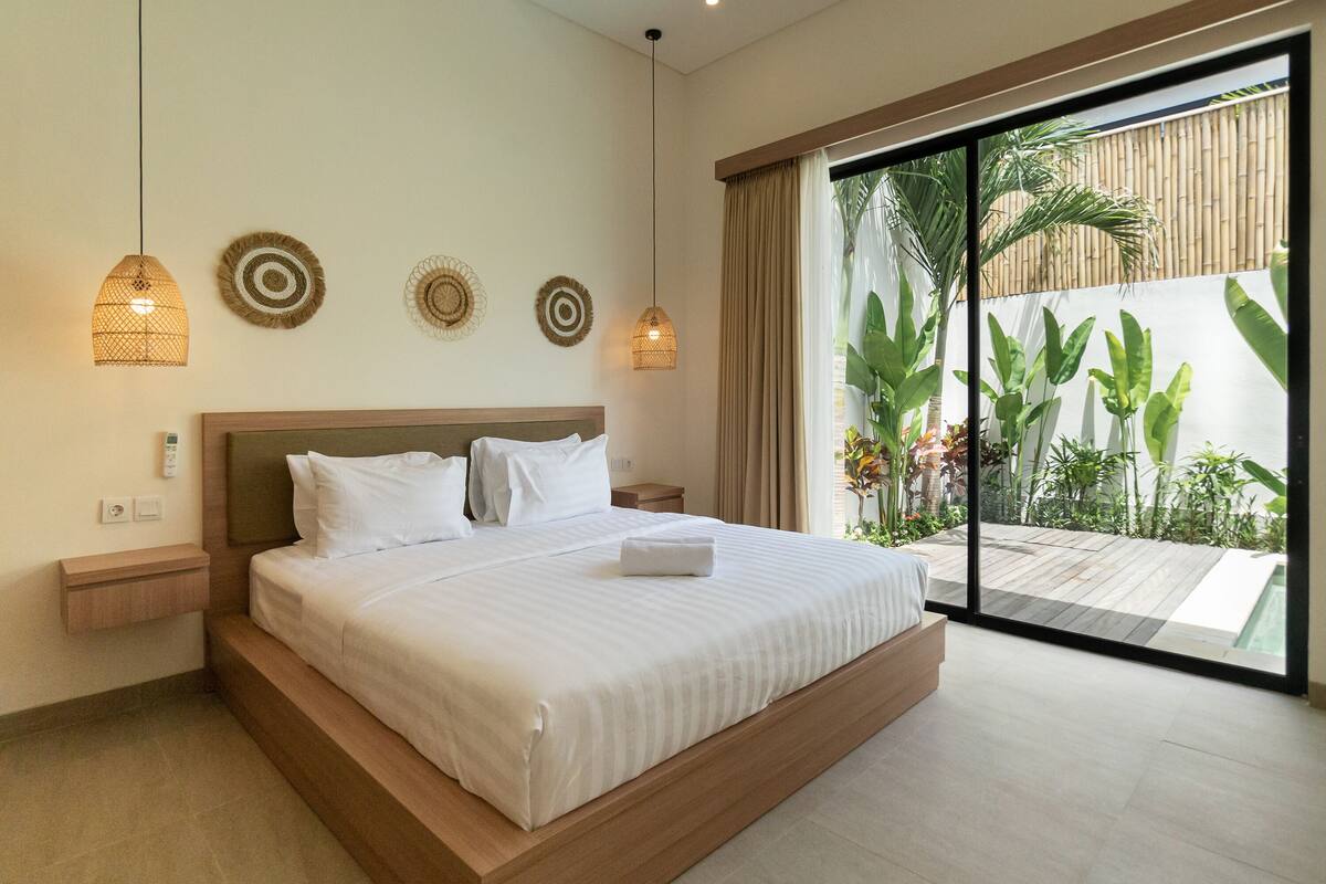 Quarto de hotel moderno com uma cama grande, cabeceira de madeira e vista para um jardim tropical através de uma porta de vidro deslizante.