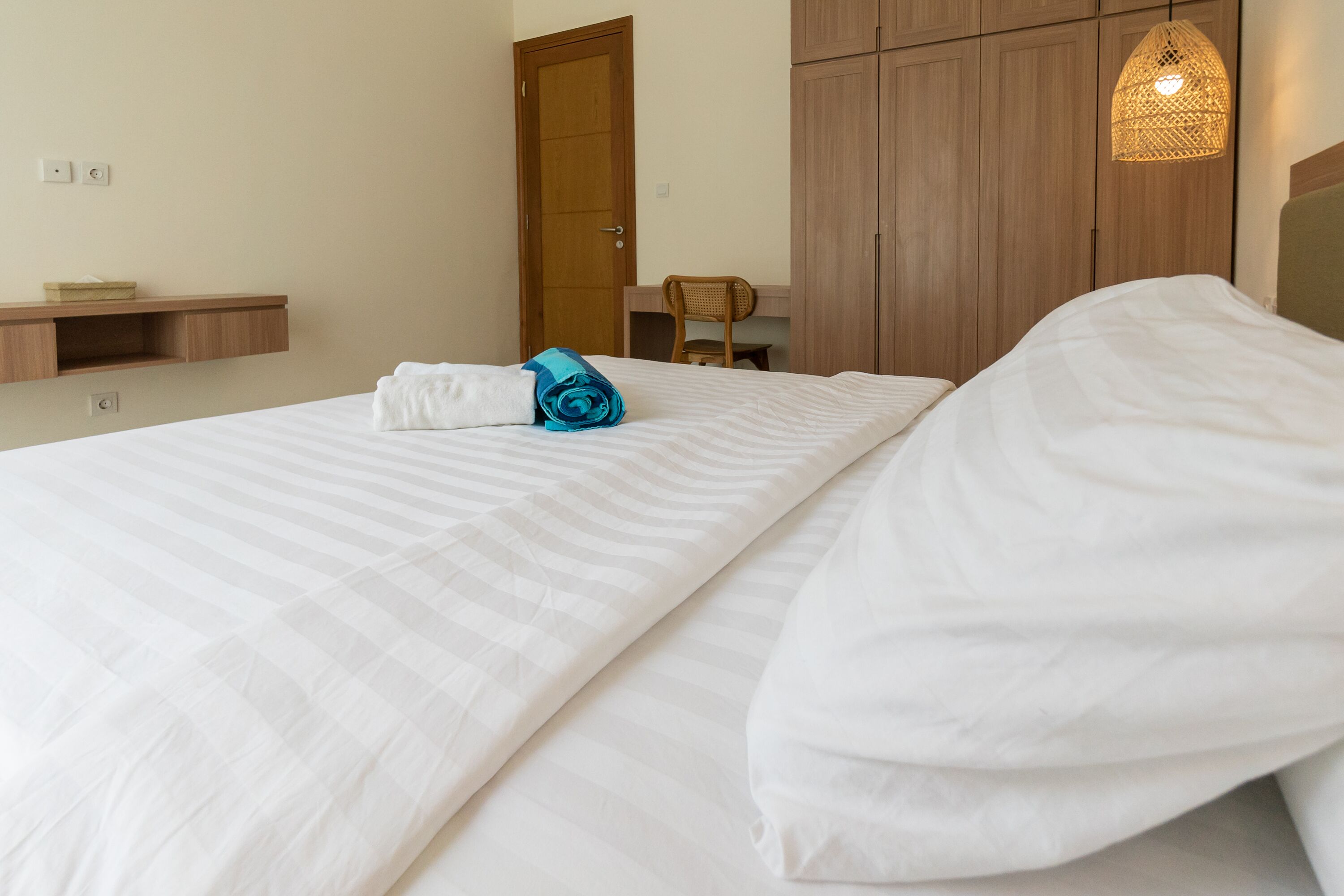 A neatly made bed with white linens and a rolled towel, a wooden wardrobe, a small wooden desk with a chair, and a hanging light fixture.