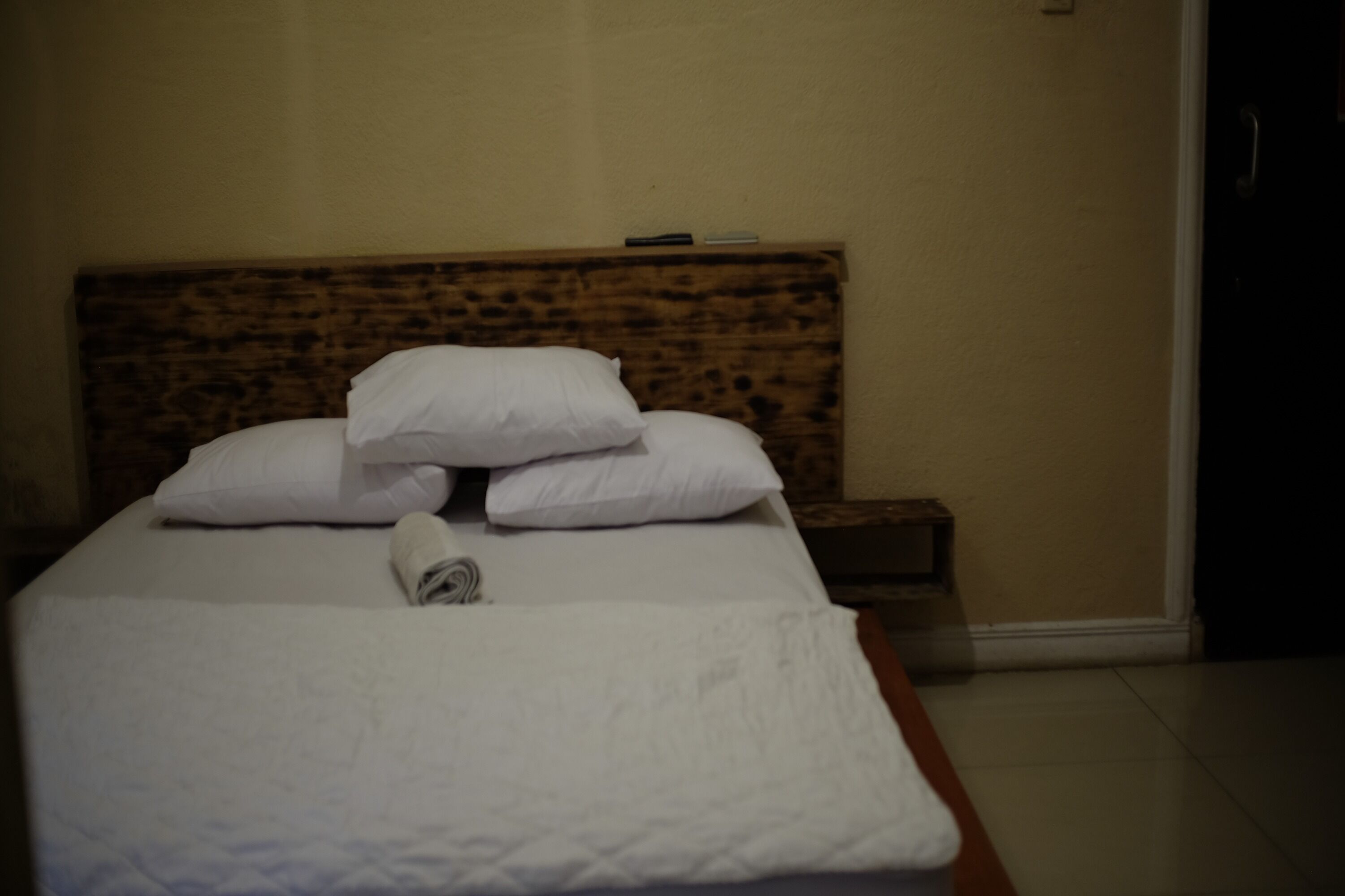 A neatly made bed with white linens and pillows, a wooden headboard, and a small bedside table with a remote control.