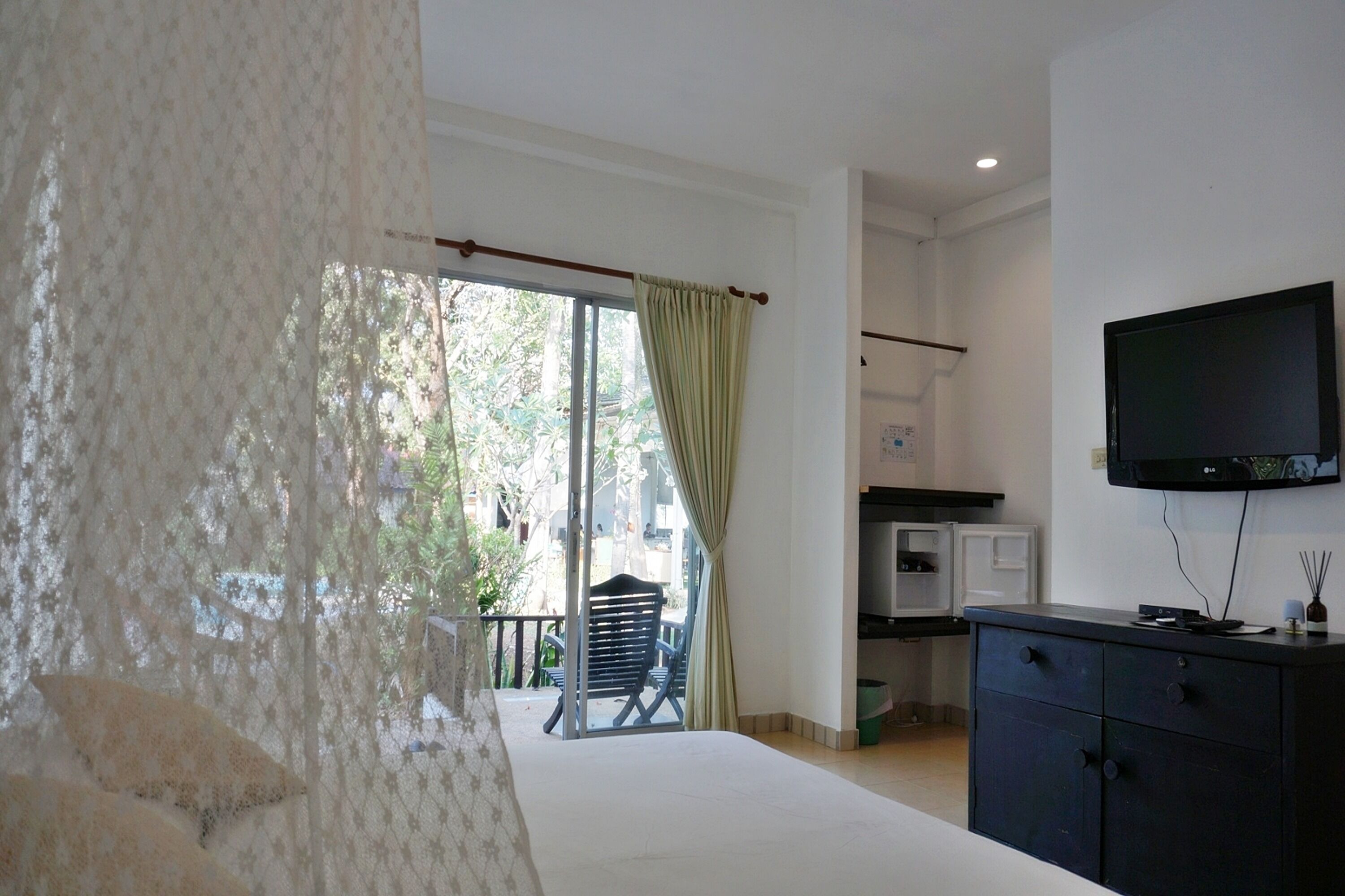 A room with a bed, a TV mounted on the wall, a small kitchenette, and a balcony with a chair.