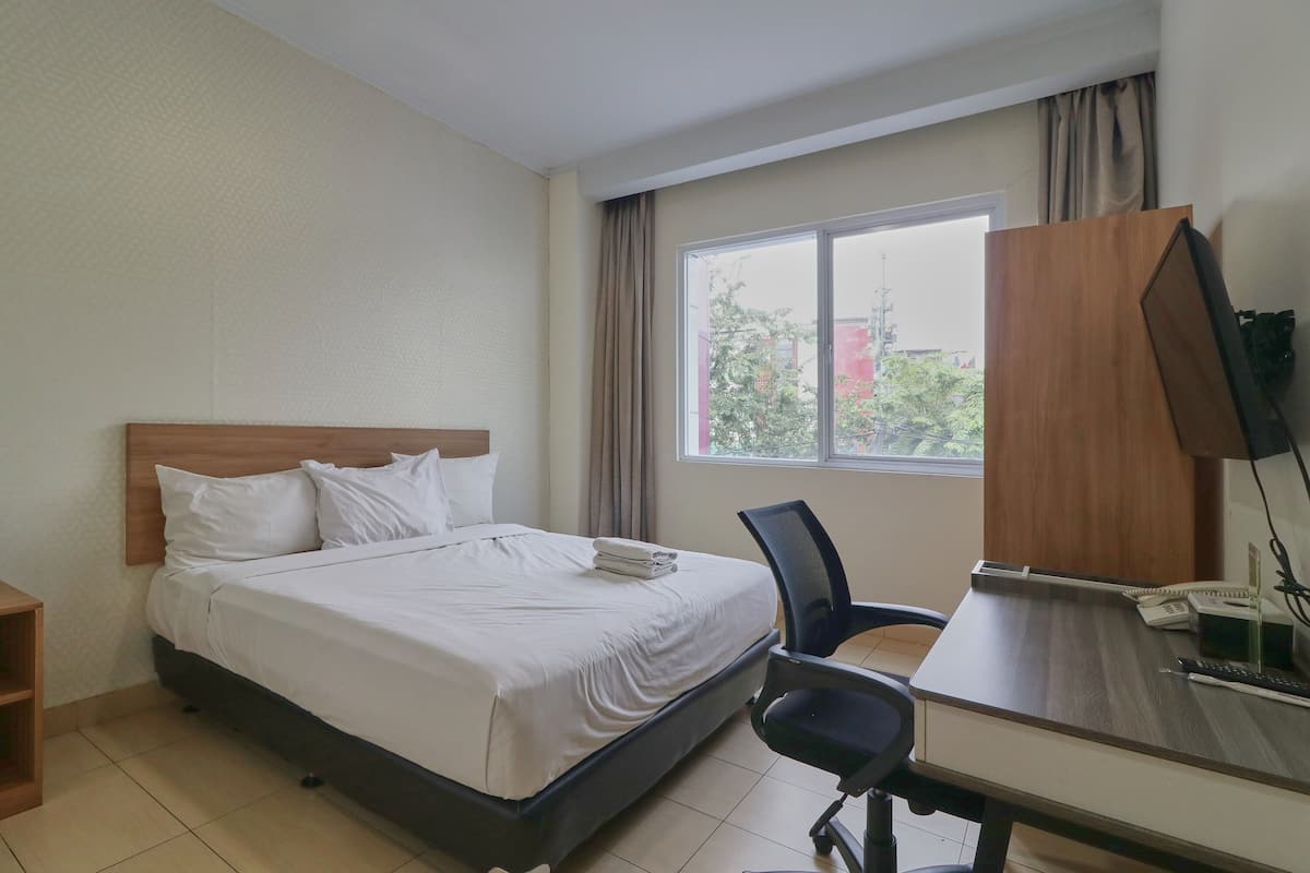 A hotel room with a bed, a desk, a chair, a TV, and a window with a view of buildings and trees.