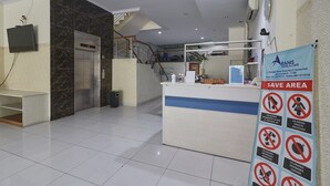 Zona de recepción de hotel con mostrador de recepción, un televisor montado en la pared, un ascensor y un cartel de "SAVE AREA" con pautas de salud y seguridad.
