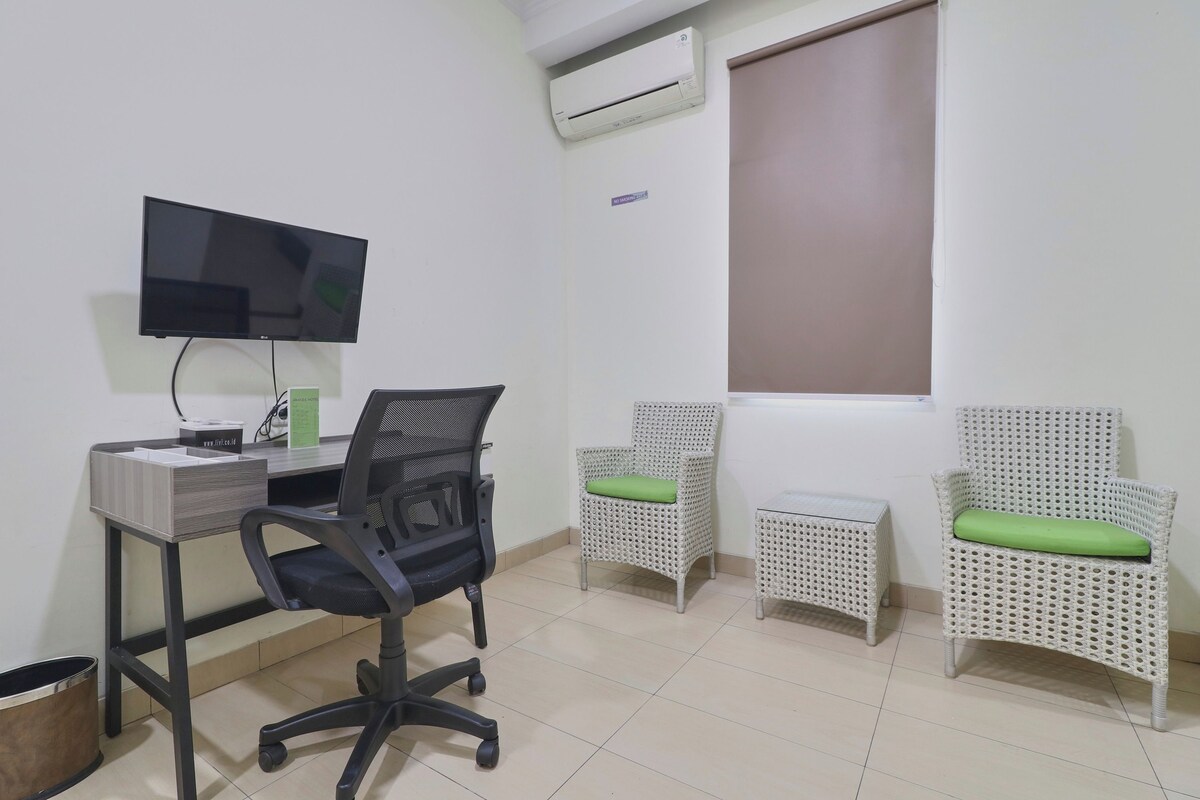A room with a desk, a black office chair, two wicker chairs with green cushions, a television mounted on the wall, and an air conditioning unit.