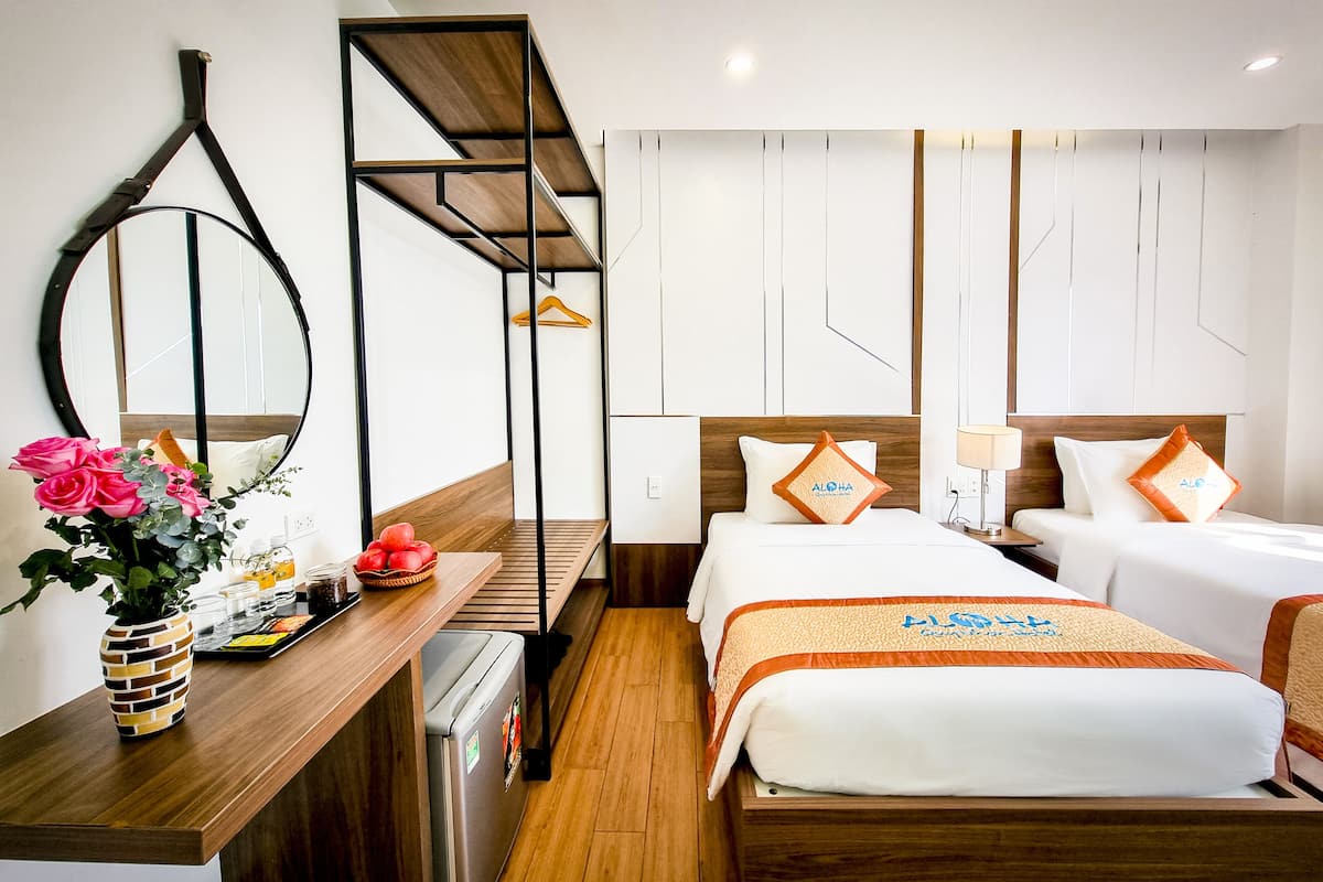 A hotel room with a wooden desk, a mirror, a bed with a white and orange bedspread, and a wall-mounted shelf with a hanging garment.