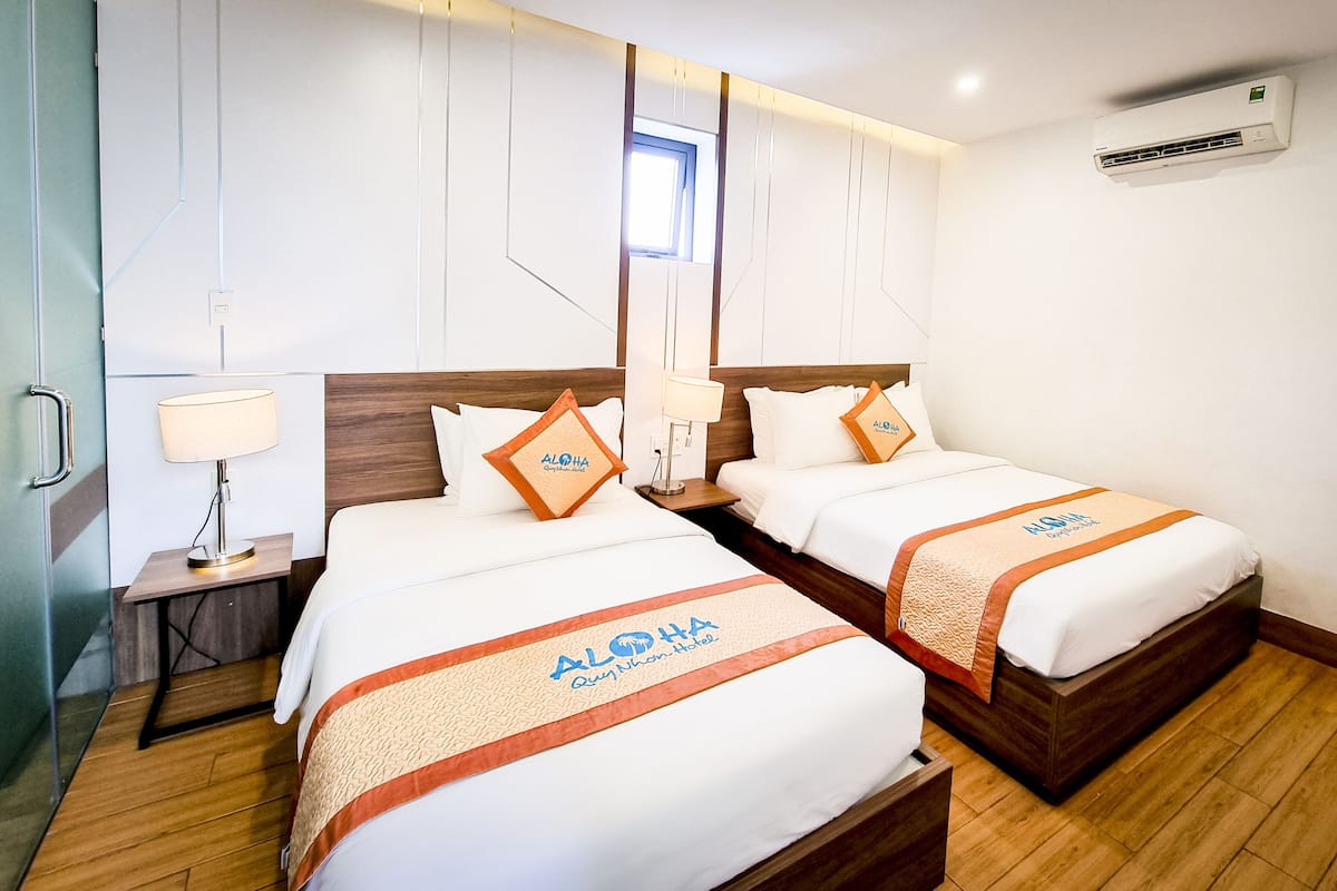 A hotel room with two beds, each with a white bedspread and an orange and white blanket. There is a nightstand with a lamp, a window with curtains, and an air conditioning unit on the wall.