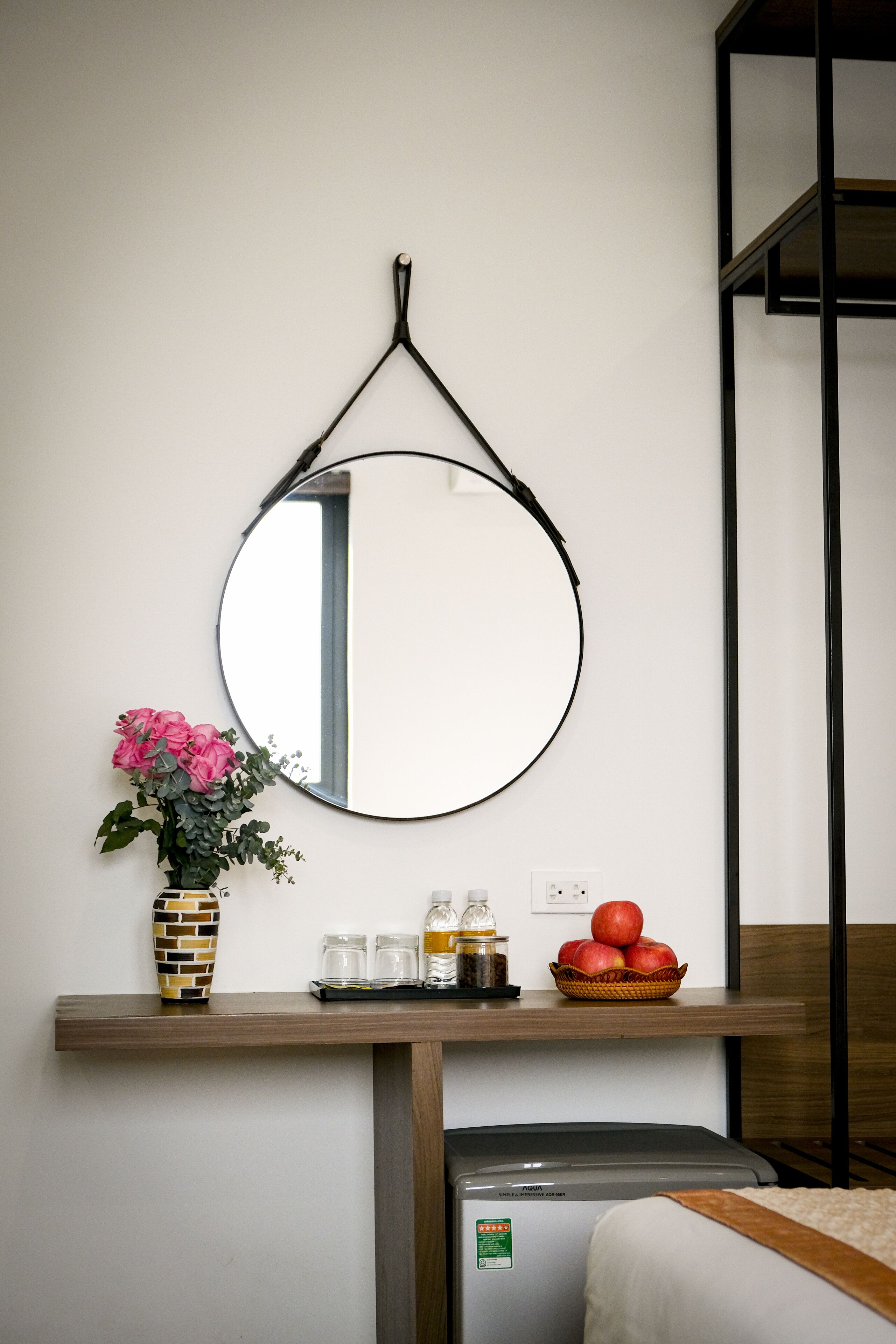 A bathroom with a round mirror, a vase with pink flowers, a basket of apples, and a small appliance.