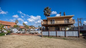 A two-story wooden building with a balcony, surrounded by a corrugated metal fence, palm trees, and a clear blue sky. - Rosarito Beach Rooms #2 (Rosarito)