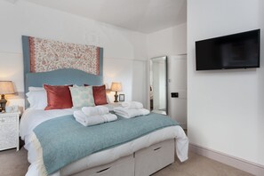 A neatly made bed with a blue headboard and white bedding, a flat-screen TV mounted on the wall, and a bedside table with lamps.