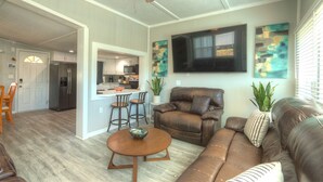 A spacious living room with a brown leather sofa, a wooden coffee table, and a ceiling fan. A kitchen is visible in the background with a refrigerator and a dining area with chairs. - 4 Bedroom Farmhouse On The River 2 (Spring Hill)
