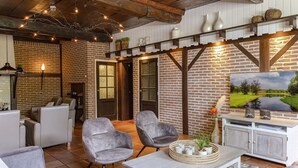 A cozy indoor space with brick walls, wooden beams, and a fireplace. There are two grey armchairs, a white table, and a television mounted on the wall. - Familieboerderij by Interhome (Losser)