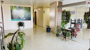 A spacious lobby with a tiled floor, potted plants, a large poster, and a seating area with chairs and tables.
