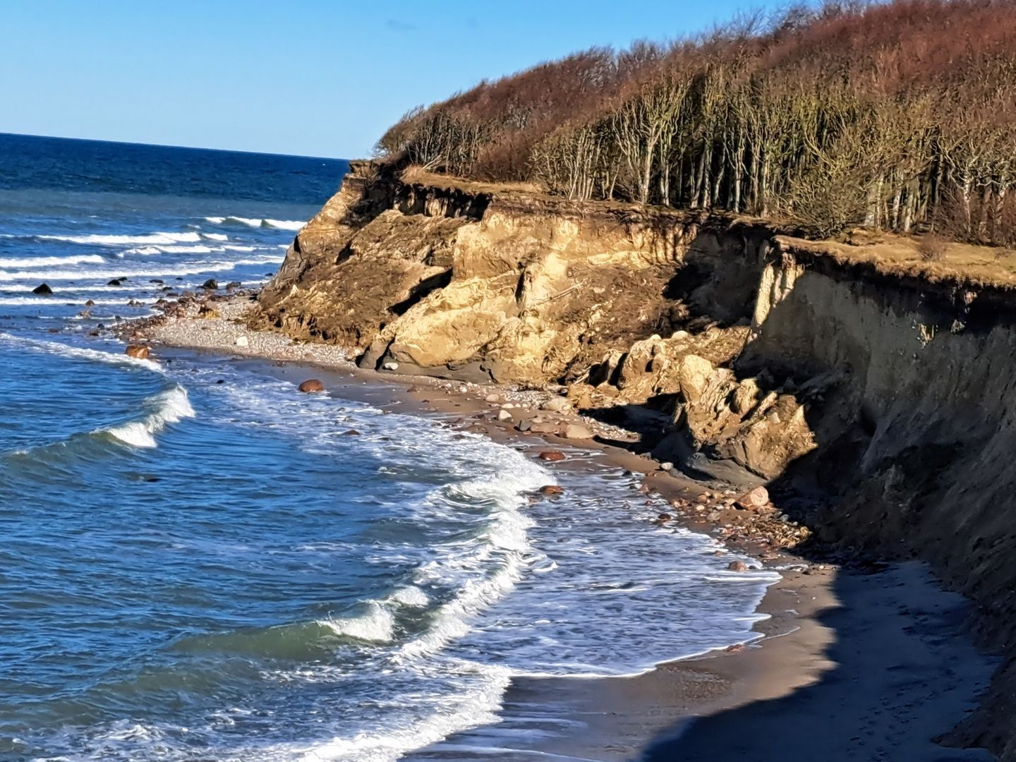 Une côte rocheuse où les vagues se brisent sur le rivage, une falaise à la végétation clairsemée et un ciel dégagé.