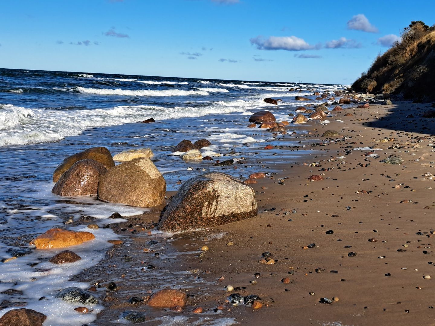 Ein Strand mit großen Felsen und Kieselsteinen, wo Wellen brechen, und ein klarer Himmel darüber liegt.