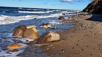 Ein Strand mit großen Felsen und Kieselsteinen, wo Wellen brechen, und ein klarer Himmel darüber liegt.