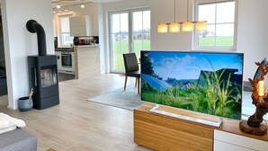 A modern living room with a large flat-screen TV, a fireplace, and a kitchen in the background.