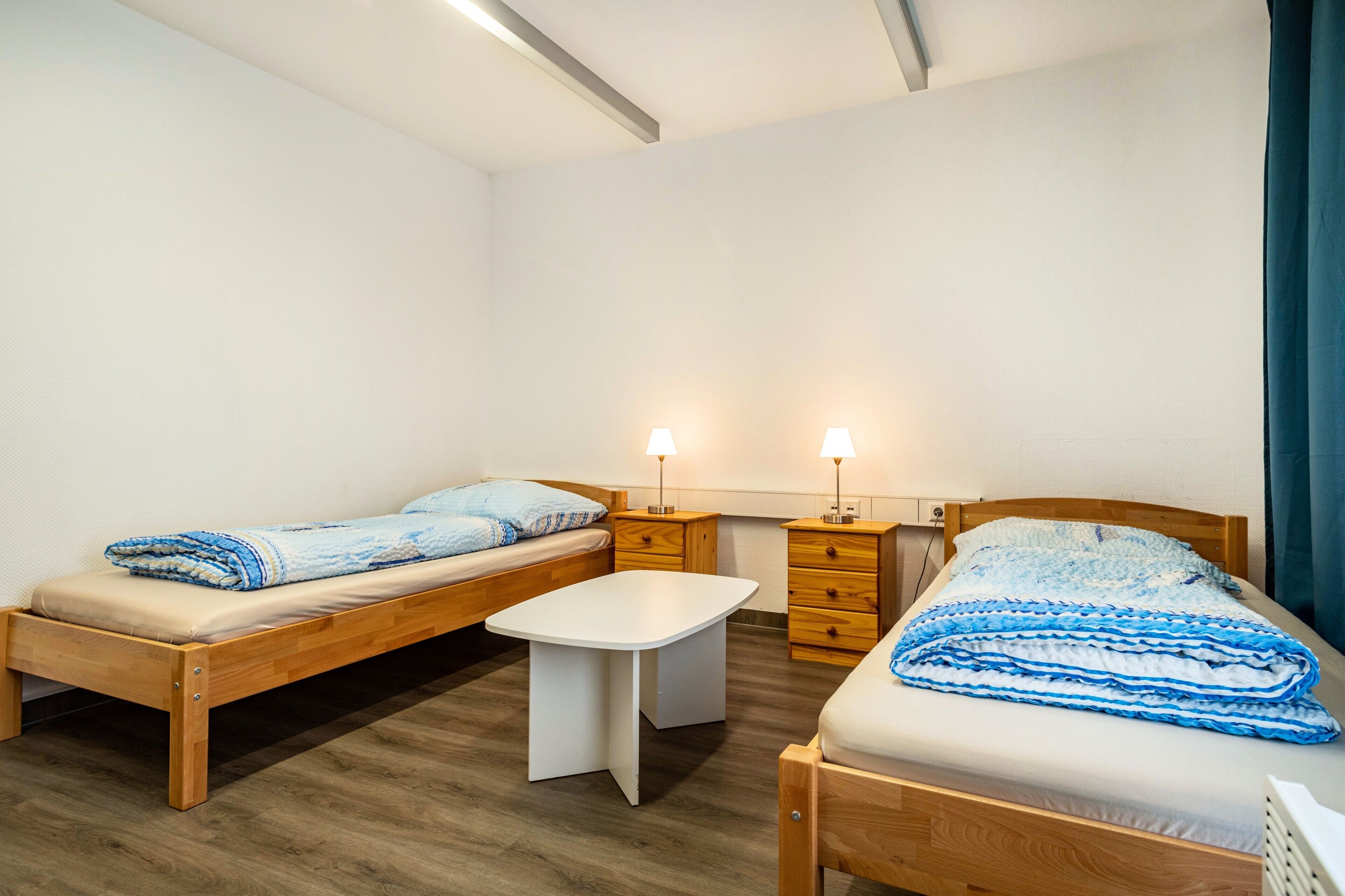 A small, well-lit room with two single beds, a small table, and two bedside tables with lamps.