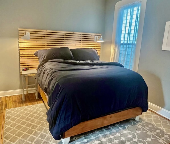 A bedroom with a bed, a bedside table, a lamp, a window, and a patterned rug.