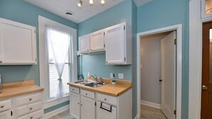 Cocina compacta con armarios blancos, encimera de madera y ventana con cortinas de gasa.