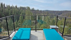 Uma varanda com duas áreas de estar estofadas em azul, guarda-corpo de vidro e vista para uma paisagem arborizada.
