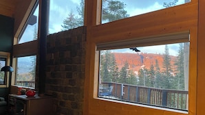 A cozy cabin interior with a stone fireplace, a wooden deck, and a large window offering a view of a forested mountain. - Sunrise Kirkwood Chalet with Red Cliffs View (Kirkwood)