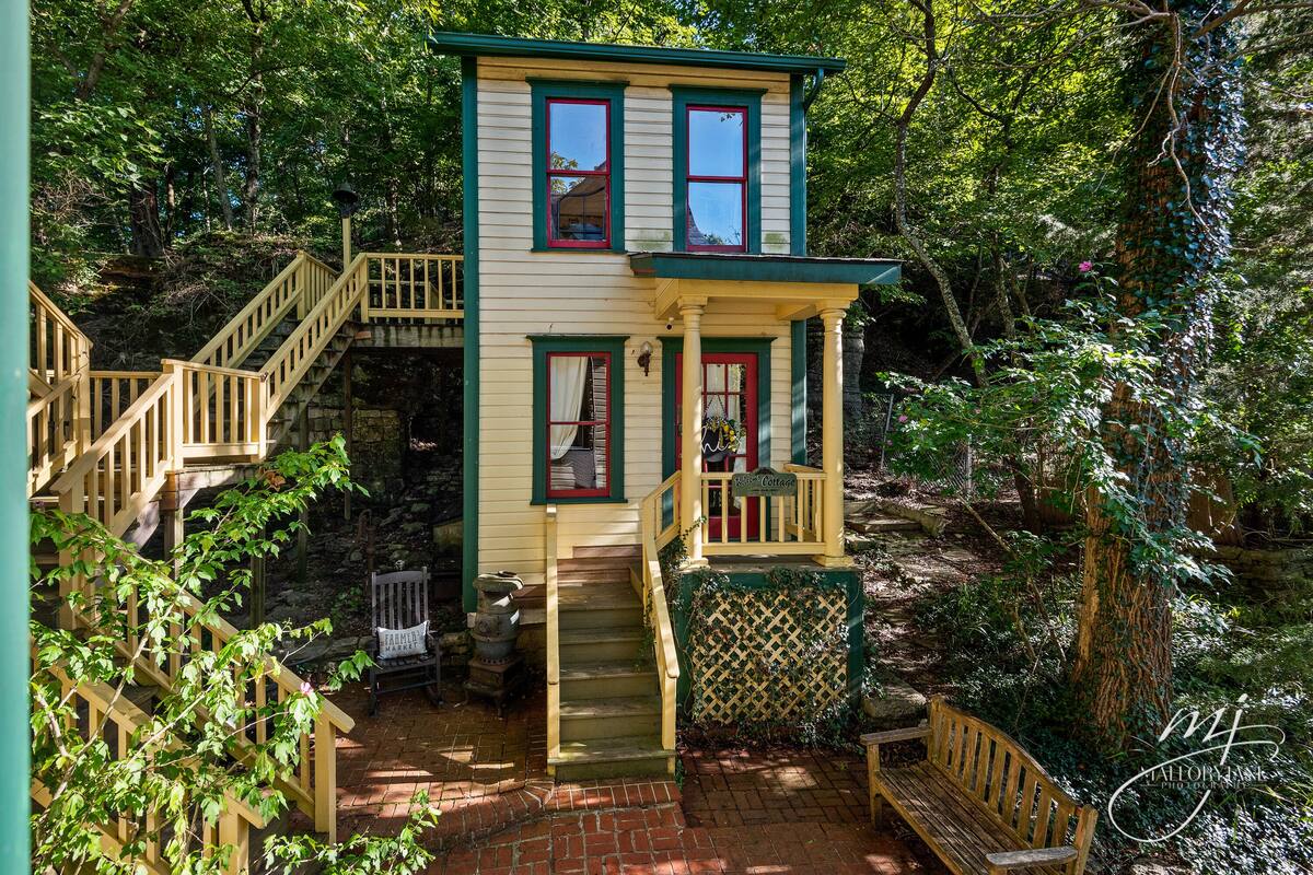 A two-story house with a wooden staircase leading to a porch, surrounded by lush greenery.
