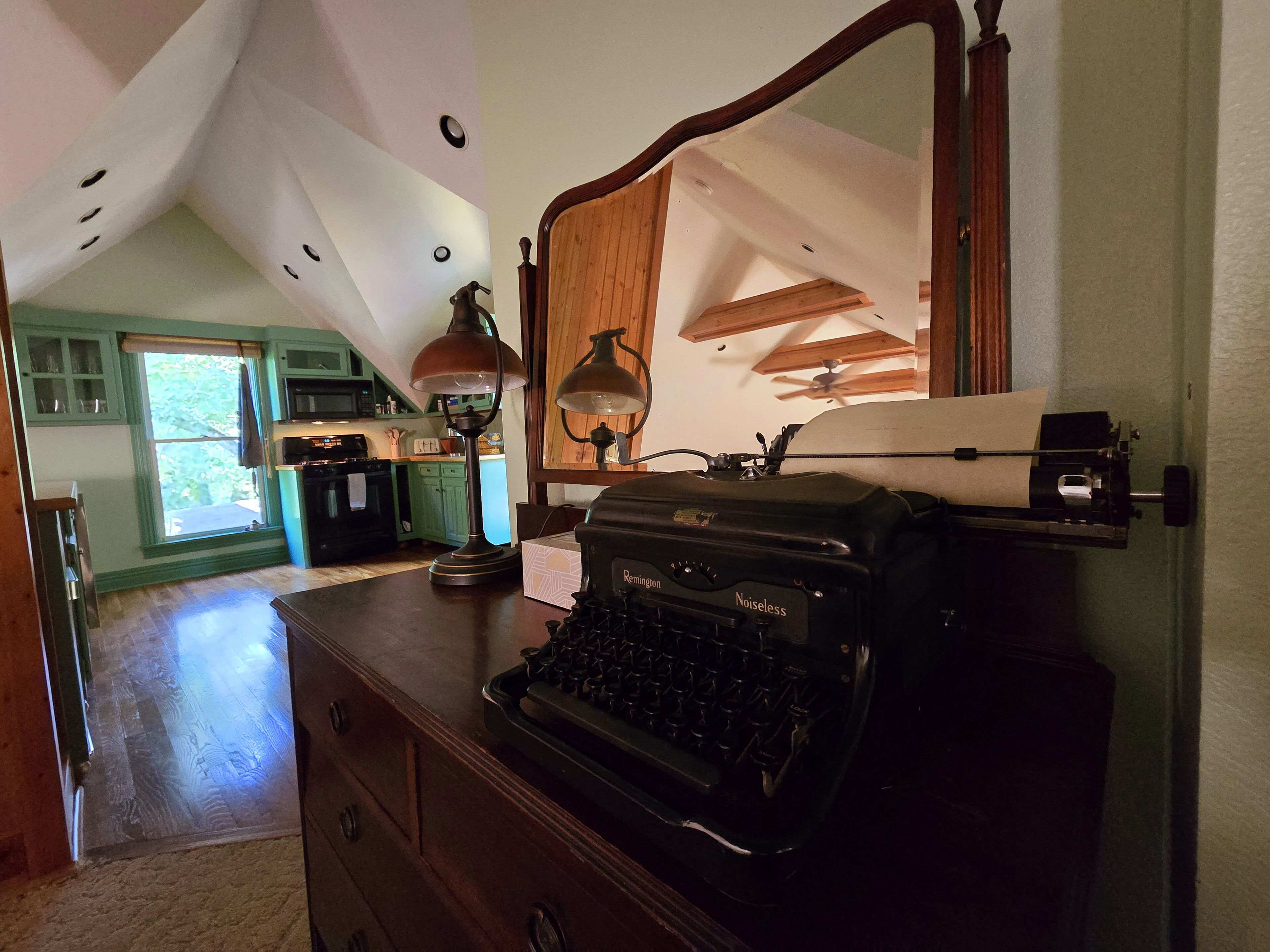 A room with a vintage typewriter, a desk lamp, and a large mirror. There is a wooden ceiling with exposed beams and a view of a kitchen in the background.