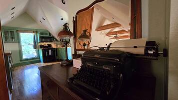 A room with a vintage typewriter, a desk lamp, and a large mirror. There is a wooden ceiling with exposed beams and a view of a kitchen in the background.