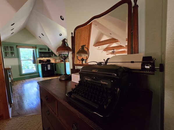 A room with a vintage typewriter, a desk lamp, and a large mirror. There is a wooden ceiling with exposed beams and a view of a kitchen in the background.