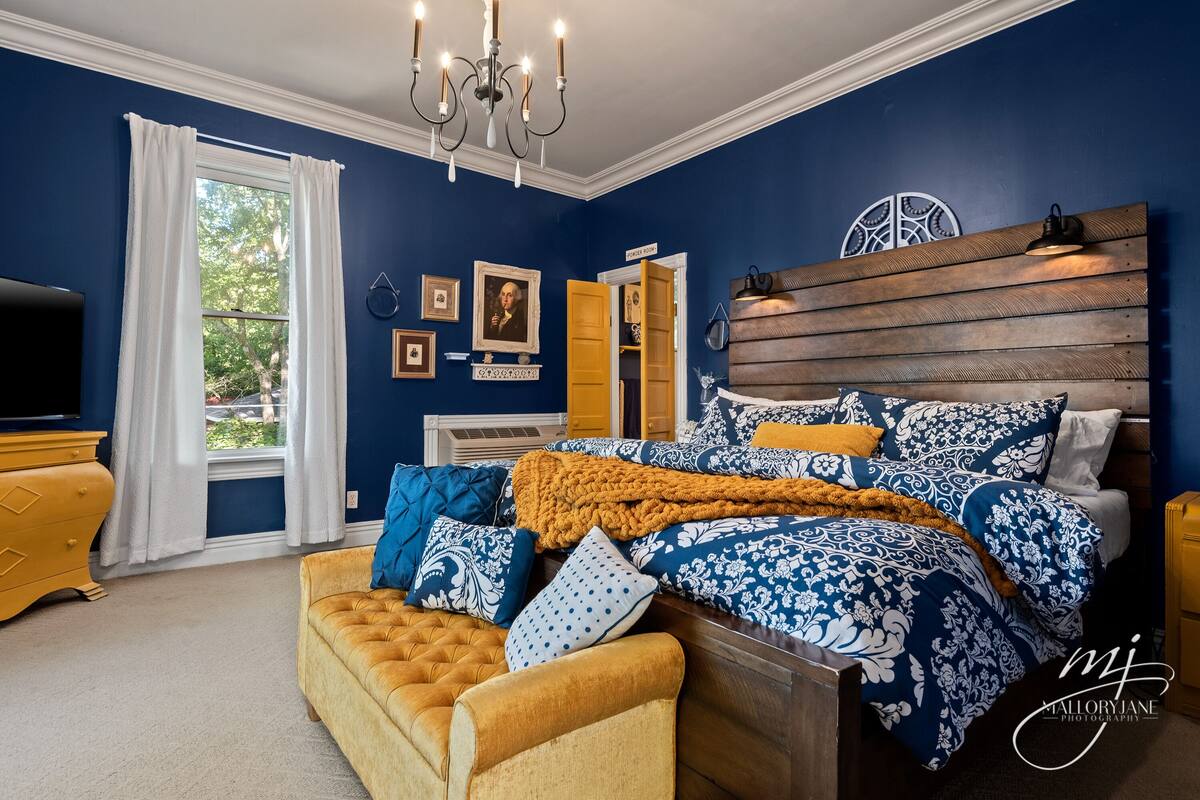 A bedroom with a large bed, a chest of drawers, a television, and a window with curtains.