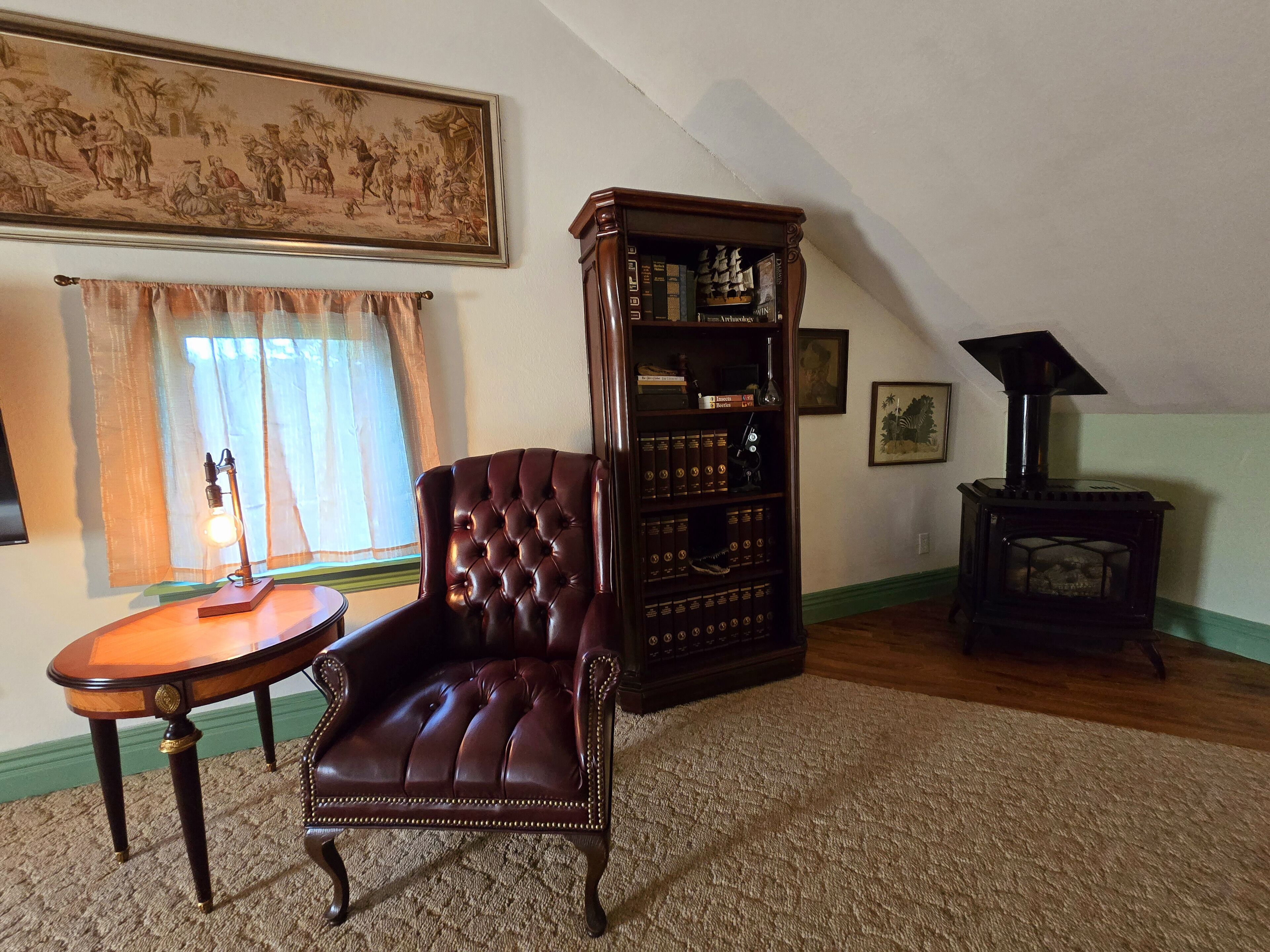 Una habitación con un sillón de cuero, una estantería de madera, una mesita con lámpara y un estufa de leña.