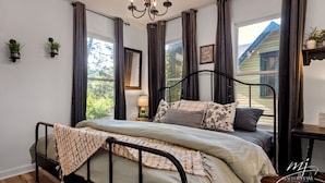 Un dormitorio con cama, lámpara de araña, ventana con cortinas, mesita de noche y estante en la pared con plantas.