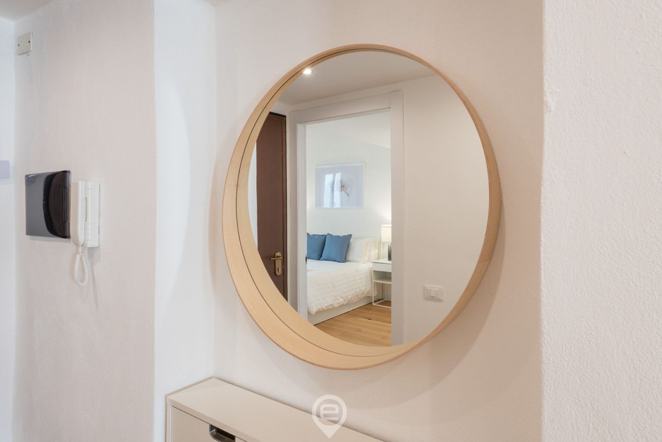 A round mirror on a wall with a visible door leading to a bedroom with a bed and a nightstand.