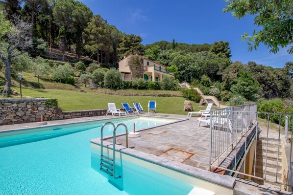 Una piscina con cubierta de piedra, rodeada por un muro de piedra y una valla de metal. Hay sillas de descanso y una escalera que lleva a una casa al fondo.