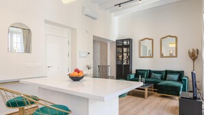 Een moderne woonkamer met een groene bank, een witte salontafel, en een keukenhoek met een wit aanrecht en een schaal fruit.