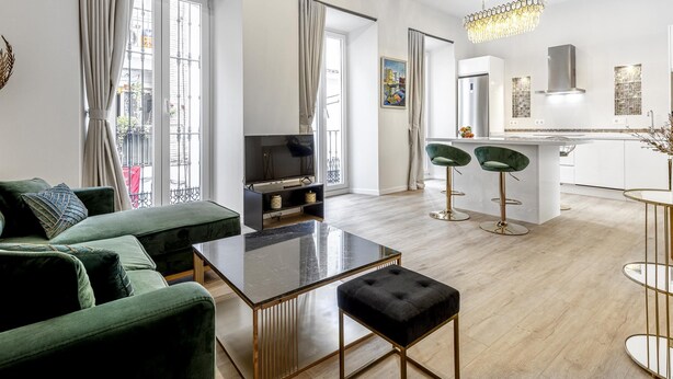 Een moderne woonkamer met een groene bank, een glazen salontafel en een televisie. Er is een keukenhoek met een bar en twee barkrukken.