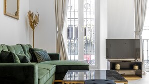 Een moderne woonkamer met een groene modulaire bank, een glazen salontafel, een zwart voetenbankje, een flatscreen-tv op een zwarte standaard en een groot raam met gordijnen.